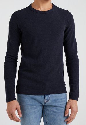 Mann trägt einen eng anliegenden, marineblauen, langärmligen Strickpullover und hellblaue Jeans, steht vor einem einfarbigen Hintergrund.