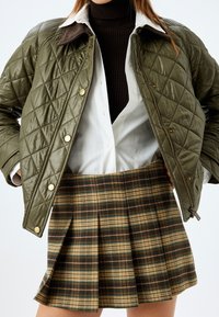Chaqueta acolchada verde con botones dorados, sobre una camisa blanca de botones y un cuello de tortuga marrón, combinada con una falda con pliegues de cuadros en tonos beige y oscuros.
