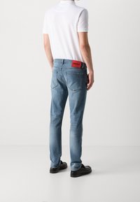 Vaqueros de mezclilla azul claro con un corte ajustado, que cuentan con dos bolsillos traseros y un parche de logo rojo en la cintura. El calzado es negro con un diseño texturizado.