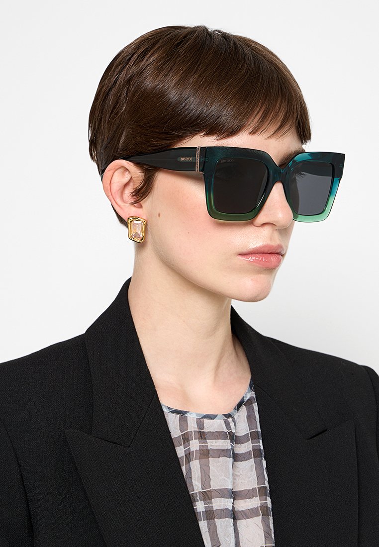Lunettes de soleil dégradé noir et vert avec de grandes lentilles carrées, arborant une monture élégante et des accents sur les côtés. Portées avec un blazer sombre et un haut à motifs.