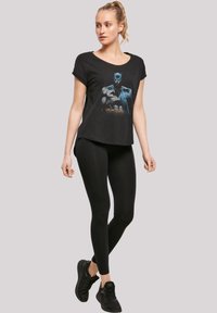 Svart t-skjorte med Batman-påtrykk, rund hals, korte ermer, kombinert med svarte leggings og svarte joggesko. Jevn tekstur gjennom hele plagget.