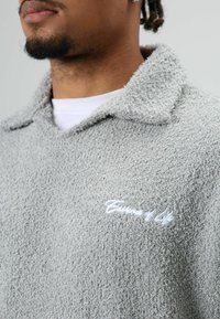 Grijze pullover met textuur en een zachte, pluizige afwerking. Heeft een kraag en een wit geborduurd merkembleem op de linkerborst.