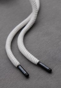 Bílé pletené tkaničky na boty s černými plastovými konci, s tištěným brandingem na koncích, ležící na strukturovaném šedém povrchu.