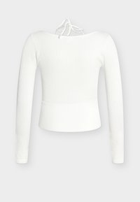 Top alb cu mâneci lungi, croșetat cu dungi, de lungime scurtă, cu decolteu barcă și închidere cu fundă în spate, pe un fundal gri deschis.