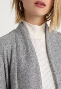 Šedý vlněný plášť s jemnou strukturou, doplněný světle béžovým žebrovaným rolákem. Ozdoben velkými stříbrnými kruhovými náušnicemi.