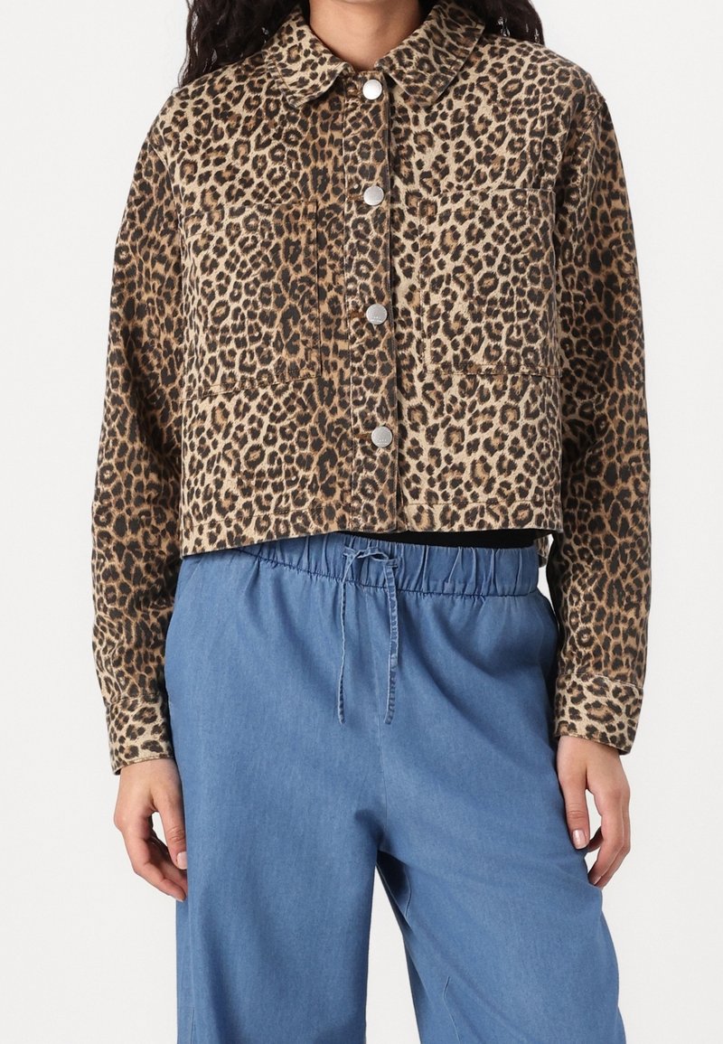 Femme portant une veste courte boutonnée à imprimé léopard et un pantalon bleu ample à cordon sur fond blanc.