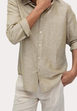 Man draagt een beige overhemd met verticale strepen met opgerolde mouwen en lichtbeige broek, de ene hand raakt de kin aan en de andere zit in de zak.