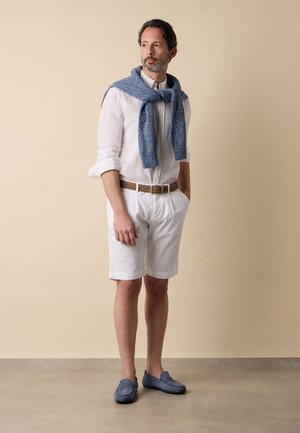 Mann trägt weißes langärmliges Hemd mit hochgekrempelten Ärmeln, weiße Shorts, braunen Gürtel, blauen Pullover über den Schultern und blaue Mokassins, stehend.