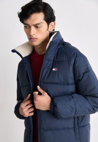 Joven ajustando una chaqueta acolchada azul marino con forro de felpa blanca, vistiendo una camiseta roja debajo, mirando hacia abajo y hacia un lado.
