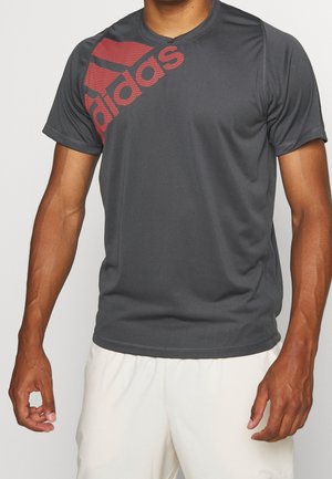 Dunkelgraues Adidas Kurzarmshirt mit großem rotem Logo auf der Schulter, getragen von einer Person mit hellbeigen Shorts, Arme entspannt.
