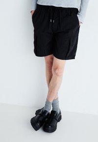 Shorts cargo noirs avec une taille élastique, dotés de poches latérales et d'une texture légère. Associés à des sabots noirs et des chaussettes grises.