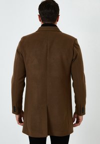 Abrigo de lana marrón con un diseño estructurado, que cuenta con un cuello con muescas, una abertura central y acentos de botones negros en las mangas.