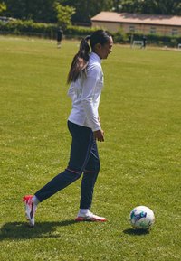 Fehér, hosszú ujjú sportpulóver, sötétkék nadrág rózsaszín részletekkel és fehér focicipő, focizik egy mintás labdával a zöld fűben.