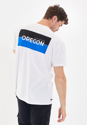Valkoinen puuvillapaita, jossa on musta ja sininen grafiikka selässä, jossa lukee "OREGON" ja "CITY OF PORTLAND". Yksinkertainen lyhythihainen muotoilu.
