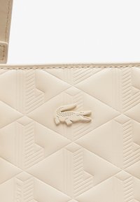 Sac à main beige avec un motif géométrique texturé. Présente un logo crocodile blanc proéminent. Le matériau semble lisse et synthétique.