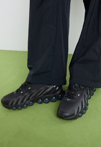 Schwarze Sportschuhe mit strukturiertem Obermaterial, geschwungenem Design und runden blauen Akzenten an der Sohle. Getragen mit lockeren schwarzen Hosen.