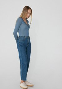 Světle modrý top s dlouhým rukávem a zvlněným výstřihem, kombinovaný s modrými džíny s vysokým pasem a béžovými teniskami.