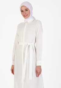 White long-sleeve dress with a shirt collar and a matching tie belt. Soft fabric with buttoned cuffs and a straight-cut design.