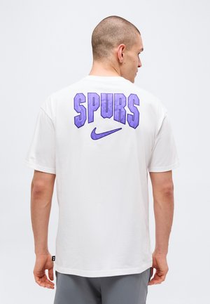 Homme aux cheveux courts portant un t-shirt blanc avec le texte violet "SPURS" et le logo Nike au dos, debout devant un fond clair uni.