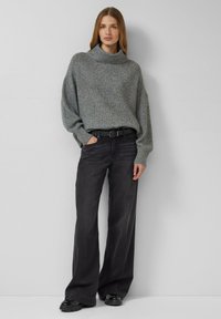 Grijze gebreide trui met een hoge hals, losse mouwen en een gestructureerd patroon, gecombineerd met zwarte wijde jeans met een hoge taille en zwarte schoenen.