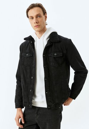 Chaqueta de mezclilla negra con cuello de sherpa, que cuenta con dos bolsillos en el pecho, botones plateados y un corte relajado, superpuesta sobre una sudadera blanca con capucha.