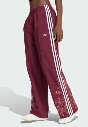 Personne portant un pantalon de jogging Adidas bordeaux avec des bandes blanches et une broderie florale près des chevilles, associée à des chaussures blanches sans lacets.