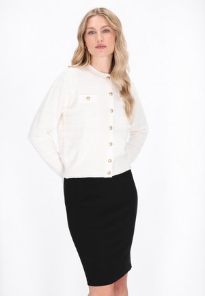 White knitted cardigan with horizontal stripes and gold buttons, paired with a fitted black skirt. Smooth texture and classic cut.