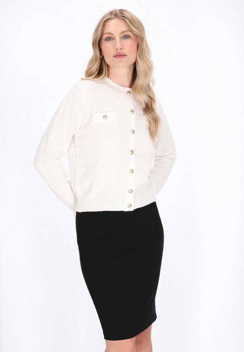 White knitted cardigan with horizontal stripes and gold buttons, paired with a fitted black skirt. Smooth texture and classic cut.