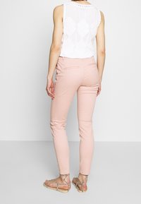 Žena v světle růžových slim-fit kalhotách, bílém topu bez rukávů s krajkovými detaily a béžových plochých sandálech na pásky, stojící na neutrálním pozadí.