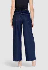 Jeans in denim scuro a vita alta e gamba larga; quattro tasche; cuciture visibili; texture liscia; tonalità di blu classica. Presenta un'etichetta in pelle sul retro.