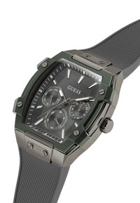 Montre Guess élégante avec un cadran texturé noir, des index heures argentés et trois sous-cadrans. Dispose d'un boîtier en métal vert et d'un bracelet en caoutchouc gris.