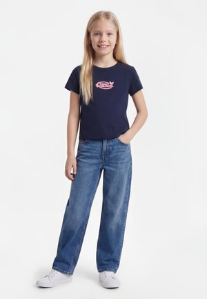 Joven chica con el pelo largo y rubio, lleva una camiseta gráfica azul marino, vaqueros azules y zapatillas blancas, está de pie con una mano en el bolsillo.