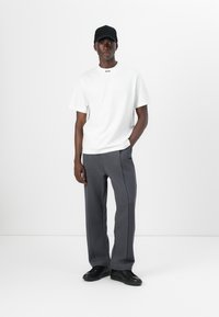 Tricou alb din bumbac cu un logo mic, purtat cu pantaloni gri cu picioare largi, având cusături laterale și adidași negri. O șapcă neagră completează look-ul.