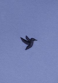 Tejido texturizado azul claro con un detallado diseño de pájaro bordado en negro situado en el lado izquierdo.