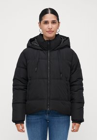 Mujer joven con cabello oscuro, vestida con una chaqueta de plumas negra con capucha y jeans azules, mirando hacia adelante contra un fondo claro y liso.