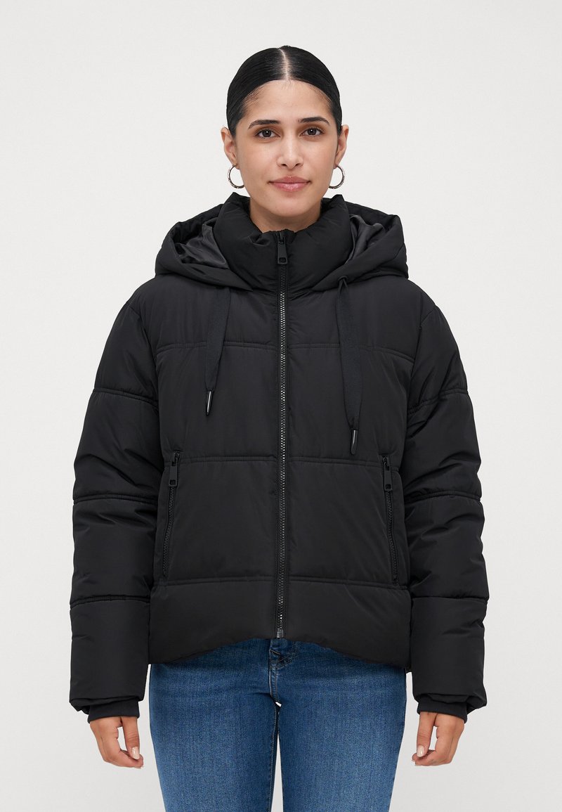 Mujer joven con cabello oscuro, vestida con una chaqueta de plumas negra con capucha y jeans azules, mirando hacia adelante contra un fondo claro y liso.
