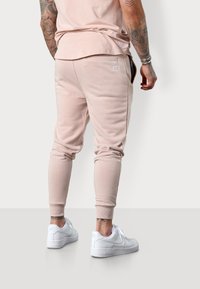 Beige Jogginghosen mit elastischen Bündchen und seitlichen Taschen, kombiniert mit weißen Sneakers. Dezente Logo-Applikation auf der Rückseite der Hose.