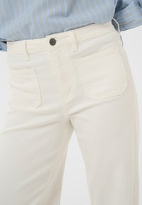 Personne portant un jean taille haute blanc cassé avec poches avant et une chemise bleu clair à fines rayures rentrée, vue de la taille à la cuisse.