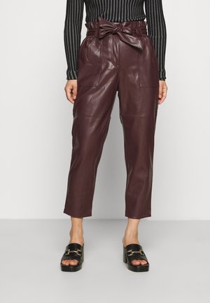 Femme portant un pantalon en cuir bordeaux taille haute style paperbag avec ceinture nouée, associé à un haut noir à manches longues rayé et des mules noires à talons.