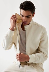 Cream bomber jacket with a high collar, zip closure, and ribbed cuffs. Smooth fabric texture; model is partially unzipping it.