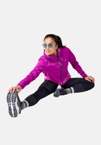 Paarse zip-up jack, zwarte leggings, reflecterende accenten en grijze sportschoenen. Persoon strekt zich uit met sportieve zonnebril.