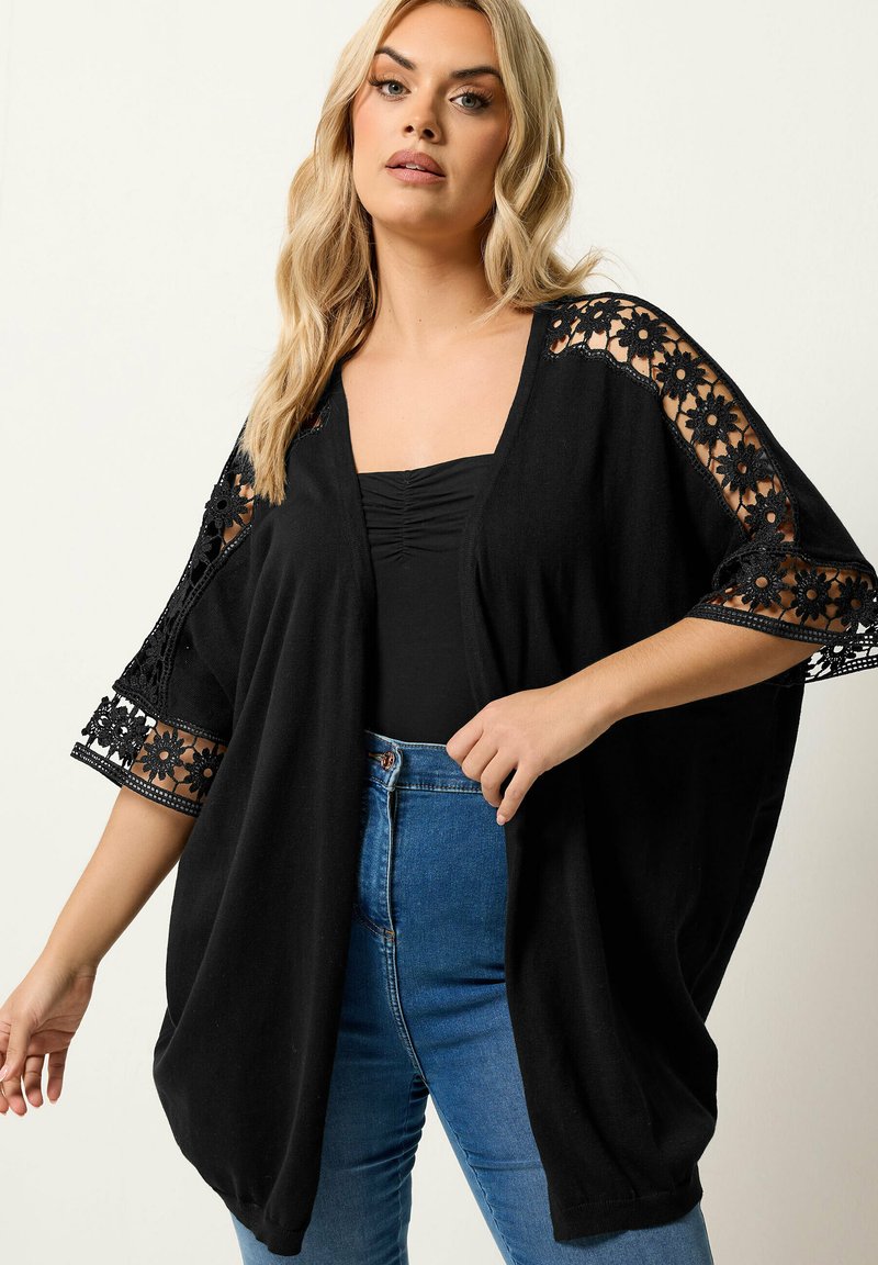 Donna bionda che indossa una maglia nera larga con maniche in pizzo floreale e jeans blu, in piedi davanti a uno sfondo chiaro e semplice.