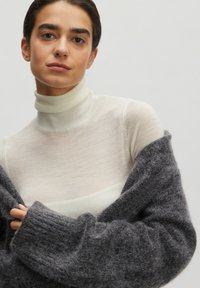 Grau, flauschiger Strickpullover über einem weißen, eng anliegenden Rollkragenpullover drapiert. Weiche Texturen, gerippte Bündchen und eine lockere Silhouette sind vorhanden.