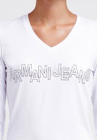 Bílé dlouhé tričko s dlouhým rukávem a výstřihem do V, s výrazným nápisem "ARMANIJEANS". Hladký bavlněný materiál, jednoduchý design.