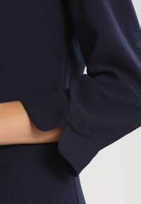 Rukáv námořnickomodré šaty, vyrobený z hladké tkaniny, s designem rozšířeného manžetu a čistými švy, s vypasovaným vzhledem.