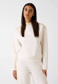 Jasno-beżowy sweatshirt wykonany z miękkiego materiału, z prążkowanymi akcentami na ramionach i okrągłym dekoltem, w komplecie z dopasowanymi spodniami.