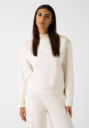 Lichtbeige sweatshirt van zacht materiaal, met geribbelde accenten op de schouders en een ronde halslijn, gecombineerd met bijpassende broek.