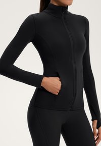 Schwarze Zip-Jacke aus dehnbarem Material. Verfügt über einen hohen Kragen und seitliche Taschen. Lange Ärmel mit eng anliegenden Bündchen, glatte Textur.