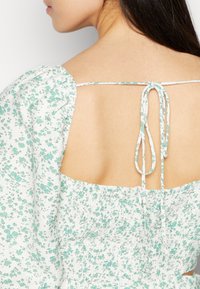 Vestido floral branco com pequenos padrões verdes, corpete elástico com franzido e delicadas alças amarradas nas costas, exibindo um design com decote baixo.
