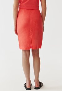 Korálová lenová tužková sukně s rozparkem vzadu, přiléhavý střih a čisté linie. Černé sandály s dekorativními akcenty dokončují outfit.